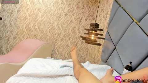 Media: Video of a nude person lying on a bed with light blue sheets, against a textured beige wall and a blue padded headboard, with a wooden pendant light hanging above.