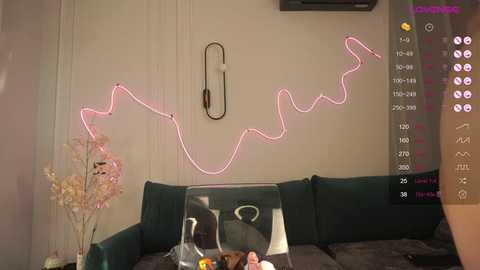 Media: Video of a modern living room with a pink neon light installation on a beige wall, a dark green couch, and a glass coffee table with a vase of pink flowers.