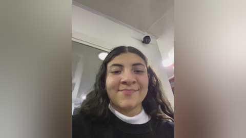 Media: Video of a young woman with long, wavy dark hair and light skin, wearing a black sweater over a white shirt, standing in a room with beige walls, a ceiling fan, and a mirror.