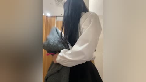 Media: Video of a person with long black hair, wearing a white long-sleeve shirt and a black skirt, hugging a grey blanket, in a beige hallway with a wooden door and a light fixture.