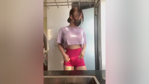 Media: Video of a young woman with light skin, wearing a purple crop top and high-waisted pink shorts, standing in a bathroom with a mirror, wearing a black face mask.