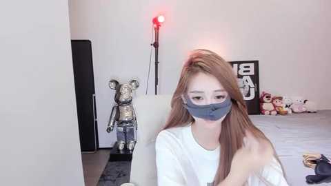 Media: Video of a young Asian woman with long, straight brown hair, wearing a white t-shirt and gray surgical mask, sitting indoors on a white chair, surrounded by a minimalist room with a teddy bear, black wall art, and red spotlight.