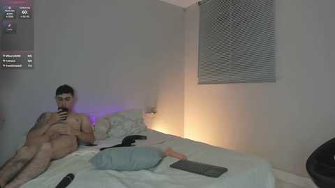 Media: Video of a nude man with tattoos, sitting on a bed in a dimly lit room, holding a smartphone, with a window with closed blinds in the background.