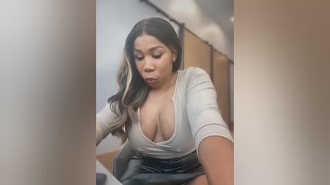 Media: Video of a young, light-skinned woman with long, wavy brown hair and large breasts, wearing a low-cut gray top and black skirt, standing in a modern, brightly-lit room.