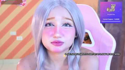 Media: Video of a young woman with light skin and long silver hair, sitting in a pink gaming chair, with a pink and orange striped wall background. Text in French on the image.