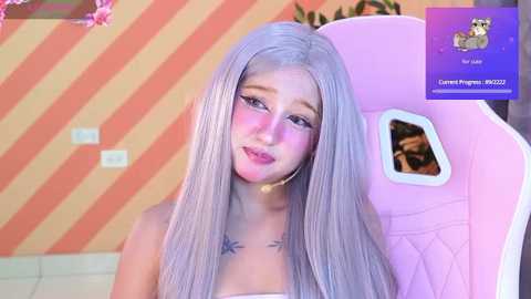 Media: Video of a young Asian woman with long, silver hair, wearing a pink gaming chair, in a room with orange and white striped wallpaper.