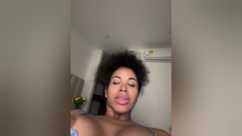 Media: A video of a young Black woman with large, curly hair, wearing a light blue bra, standing in a beige-walled room with a refrigerator and air conditioning unit in the background.