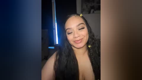 Video of a smiling, light-skinned Black woman with braids, wearing a low-cut black top, standing indoors with dim lighting.