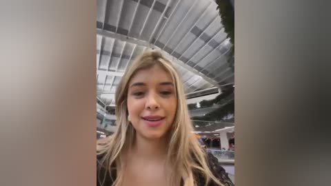 Media: Video of a young woman with long, blonde hair and fair skin, wearing a black top, smiling in a modern, indoor mall setting with a glass ceiling and greenery.