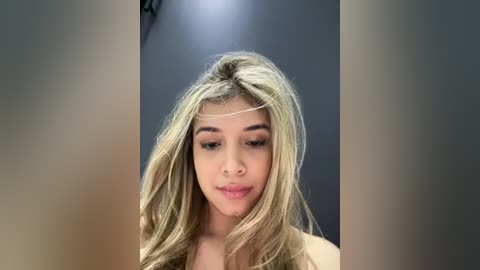 Media: Video of a young woman with long, wavy blonde hair, light skin, and subtle makeup. She gazes downwards, with a neutral expression. The background is blurred, with a gradient of dark to light gray.