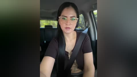 Media: Video of a young woman with long black hair, glasses, and a brown cardigan, driving a car, wearing a seatbelt. The interior is dimly lit, and the background shows greenery outside the window.