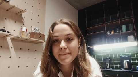 Media: Video of a young Caucasian woman with long, wavy brown hair, wearing a white blouse, standing in a modern kitchen with wooden pegboard, glass cabinets, green tiles, and a stainless steel coffee maker.