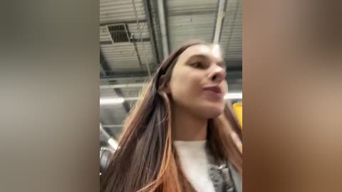 Media: A video of a young woman with long, straight brown hair and a white top, standing in a modern industrial space with exposed metal beams and pipes.