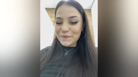 Media: A video of a young woman with long, straight dark hair, wearing a black top, silver hoop earrings, and a silver necklace. She has a serene expression, with closed eyes and a slight smile, set against a blurred indoor background.