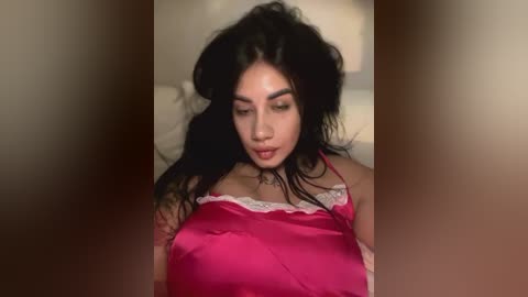 Media: Video of a young woman with long, dark hair and fair skin wearing a pink satin nightgown with white lace trim, lying on a bed with beige pillows. She appears relaxed and contemplative.