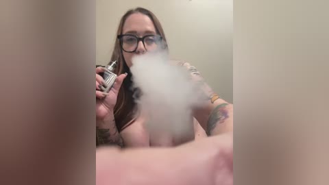 Media: Video of a topless, tattooed white woman with glasses, smoking a vape, holding a vape pen in one hand.