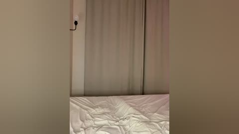 Media: A video of a minimalist bedroom with beige walls, featuring a neatly made bed with white sheets, a beige curtain, and a single white electrical outlet on the wall.