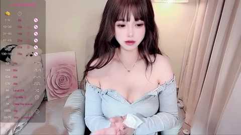 Media: Video of an East Asian woman with long, dark hair, wearing a light blue off-shoulder top, seated on a bed with a pink floral pillow, smiling gently, in a cozy, softly lit room.