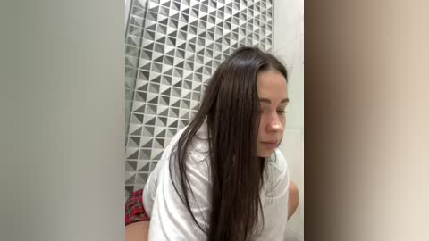 Media: Video of a young woman with long, straight, dark hair, wearing a white T-shirt and red plaid shorts, leaning against a wall with a geometric-patterned mosaic background.