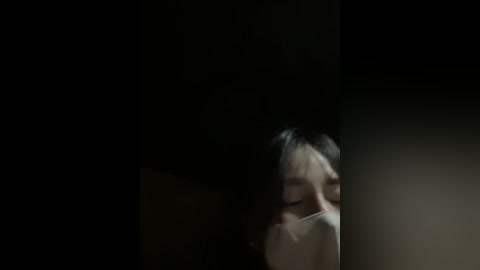 Media: A dimly-lit video showing a young woman with fair skin, short dark hair, and a white mask, her eyes closed, standing in a dark, shadowy room.