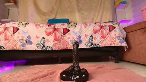 Media: Video of a black, realistic-looking sex toy on a pink rug in a bedroom with a bed covered in a butterfly-patterned duvet, beige curtains, and purple lighting.