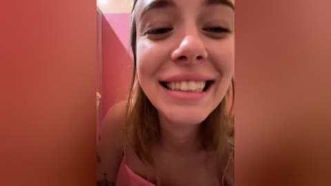 Media: A close-up video of a young, smiling Caucasian woman with light skin and straight, shoulder-length blonde hair. She wears a pink top. The background is a pink wall with a blurry door frame.