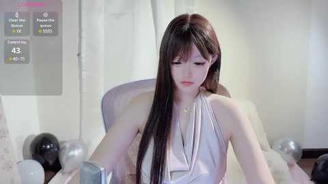 Media: Video of a fair-skinned, slender woman with long, straight brown hair, wearing a sleeveless white dress, sitting on a white chair in a modern, minimalist room.