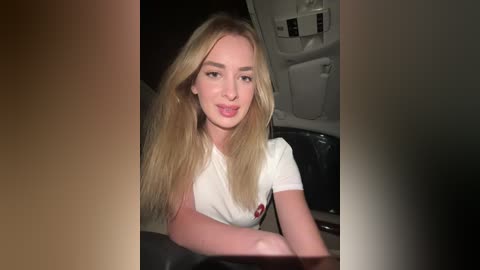 Media: A video of a blonde, fair-skinned woman with a slim physique and long hair, wearing a white t-shirt, sitting in a dimly lit bathroom with a white toilet in the background.