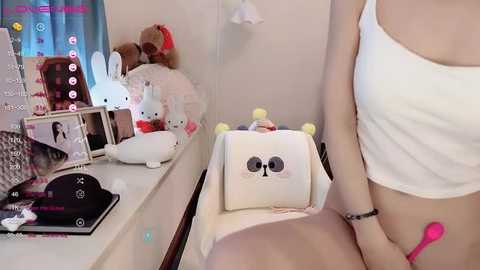 Media: Video of a light-skinned woman in a white tank top, sitting on a white chair, holding a pink vibrator, in a cluttered room with stuffed animals, framed photos, and a bed.