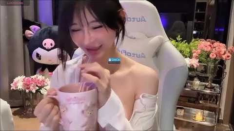 Media: A video of an Asian woman with straight black hair, wearing a white robe, drinking tea. Background features a plush toy, flowers, and a gaming chair.