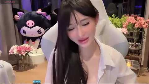 Media: Video of a young Asian woman with long black hair, wearing a white shirt, seated in a plush chair, with a plush pig behind her and floral arrangements in the background.