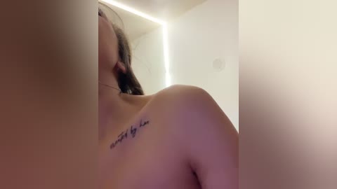 Media: Video of a topless woman with light skin and dark hair, showing a small tattoo on her shoulder reading \"Happiness is a choice.\" Background features a minimalist white room with a soft, ambient light.