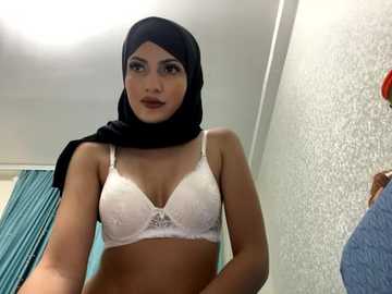 Media: Video of a young woman with light brown skin, wearing a white lace bra and black hijab, standing in a simple room with a teal curtain and textured white wall.