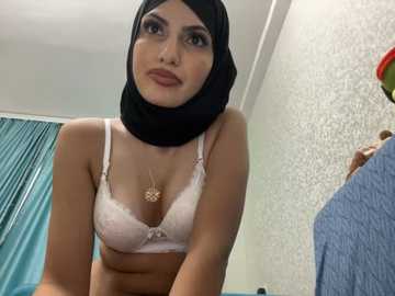 Media: Video of a light-skinned woman with a slender physique and medium-sized breasts, wearing a white lace bra and black hijab, standing indoors with a textured wall and teal curtain in the background.