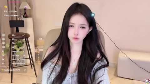 Media: A video of a young, fair-skinned woman with long, black hair, wearing a gray off-shoulder top, seated indoors with a modern, minimalistic decor background.