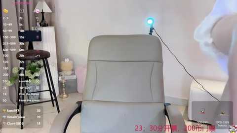 Media: Video of a beige recliner chair with a blue light emitting from a small speaker on the backrest, positioned in a modern, minimalist living room with white walls, grey curtains, and a black metal stool.