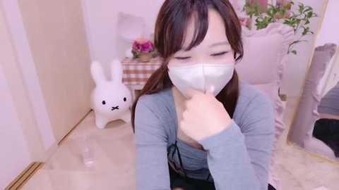 Media: Video of an East Asian woman with long brown hair, wearing a blue top, white mask, and black pants, seated indoors near a white bunny figurine and a green plant.