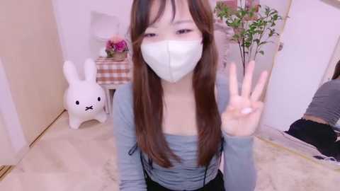 Media: Video of an East Asian woman with long brown hair, wearing a white face mask, gray top, and black pants, waving in a cozy, light-colored room with a white rabbit toy and a potted plant.