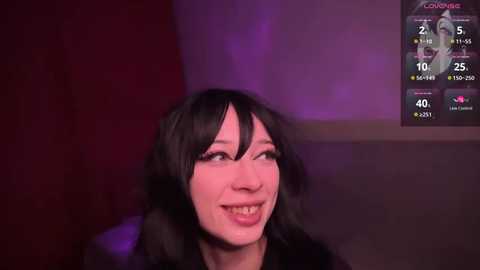 Media: Video of a smiling Asian woman with black hair and glasses, wearing a black top, in a dimly lit room with purple lighting, viewed from a webcam.