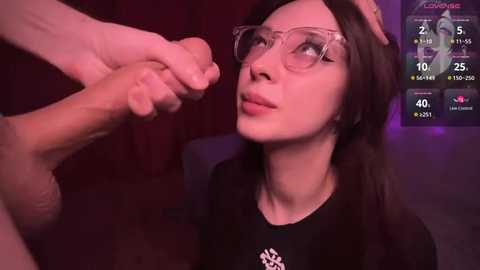 Media: Video of a young Asian woman with glasses and a black shirt, gazing at an erect penis held by a hand. Background shows a dark, dimly-lit room with digital numbers.