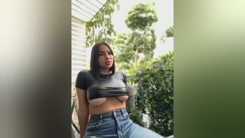 Media: A video of a young, curvy woman with medium skin tone and shoulder-length dark hair, wearing a black cropped top and high-waisted jeans, posing on a suburban porch with greenery in the background.