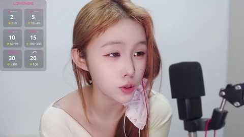 Media: A video of a young Asian woman with light skin and long, straight, reddish-brown hair, wearing a white top. She's holding a pink mask close to her mouth, possibly in a recording setup.