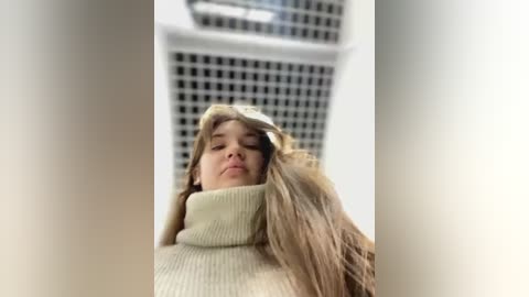 Media: A video of a young woman with long blonde hair, wearing a beige turtleneck sweater, standing in front of a grid-patterned wall, taken from a low-angle perspective.