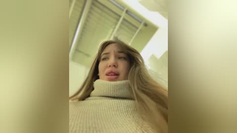 Media: Video of a woman with long brown hair, wearing a beige turtleneck sweater, captured from a low angle. She appears distressed, with her mouth slightly open, in a dimly lit room with closed blinds.