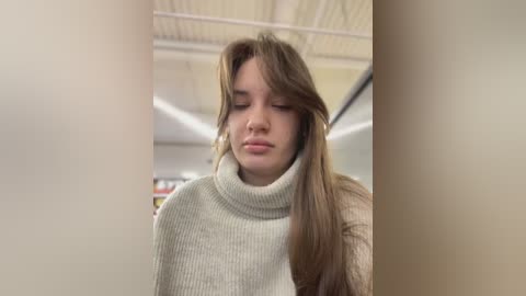 Media: Video of a young woman with long, brown hair, wearing a light gray turtleneck sweater, looking contemplative and slightly pensive, in a modern, brightly lit indoor setting with visible ceiling panels and fluorescent lighting.