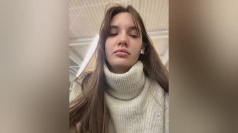 Media: Video of a young woman with long brown hair, wearing a cream-colored turtleneck sweater, looking contemplative in a fluorescent-lit room with a white ceiling.