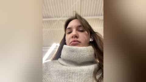 Media: A close-up video of a young woman with long brown hair, wearing a cream turtleneck sweater, lying on a white, ribbed textured ceiling. Her eyes are closed, and she appears relaxed.