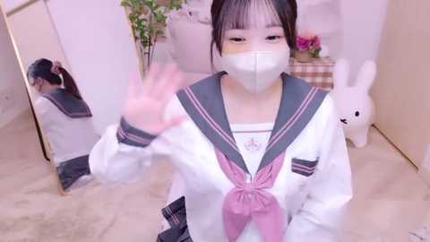 Media: Video of an East Asian woman with pale skin, dark hair, and bangs, wearing a white schoolgirl uniform with a pink scarf, mask, and bunny ears, waving in a room with soft, pastel decor and a white rabbit plush toy.