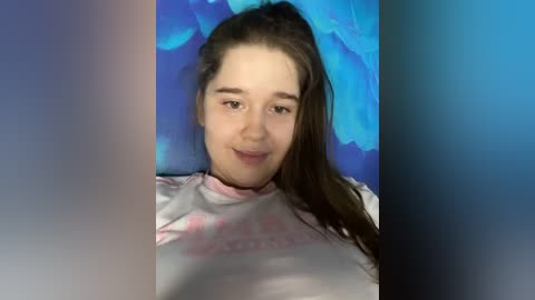 Media: Video of a young woman with light skin and long brown hair, wearing a pink and white t-shirt, smiling against a blue floral backdrop.