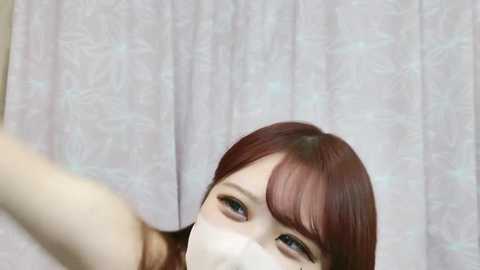 Media: Video of a fair-skinned woman with red hair, wearing a white mask, capturing a selfie with her right hand extended. She has green eyes and is indoors with a light-colored, patterned curtain in the background.